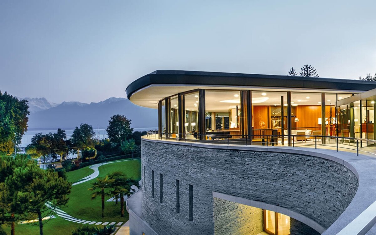 Clinique La Prairie curved stone spa building illuminated at dusk with Lake Geneva and Alpine mountains